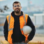 Confident male construction worker with safety vest and hard hat in a blurred outdoor setting.