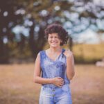 A happy woman with curly hair stands smiling in a sunny outdoor park setting.